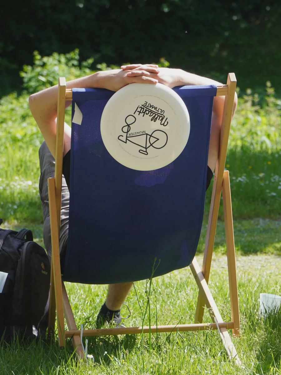 Eine Person ruht in einem blauen Liegestuhl mit Fachschaftslogo auf der Wiese.