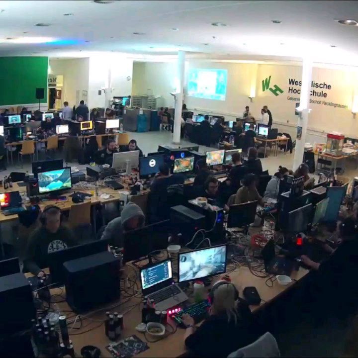 Teilnehmende sitzen an zahlreichen Computern in der abgedunkelten Mensa; im Hintergrund leuchtet eine Projektion.