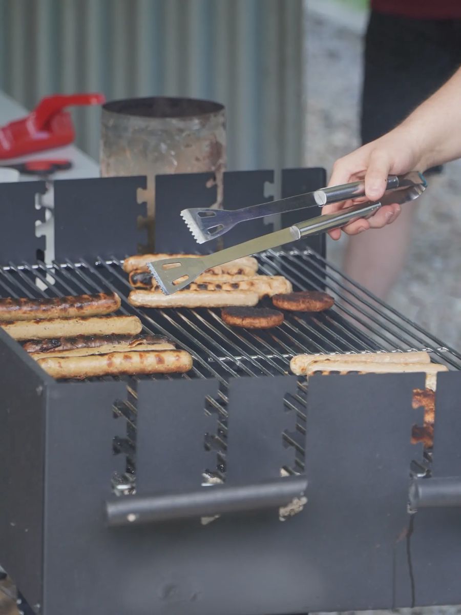 Mit einer Grillzange werden Würstchen und Fleisch auf einem Grill gewendet.