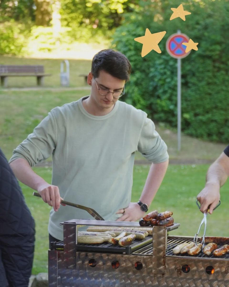 Eine Person mit Brille und hellem Pullover wendet das Grillgut; im Hintergrund stehen Tische auf der Wiese.