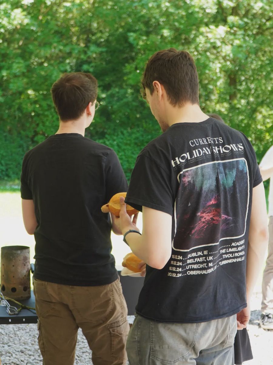 Zwei Personen stehen mit Tellern am Ausgabetisch vor einer grünen Hecke.