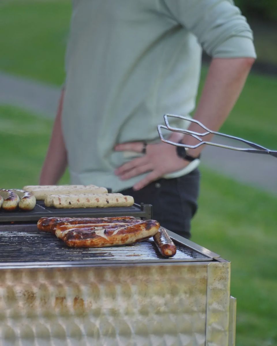 Nahaufnahme von Würstchen und Fleisch auf einem Grill neben einer Person mit Grillzange.