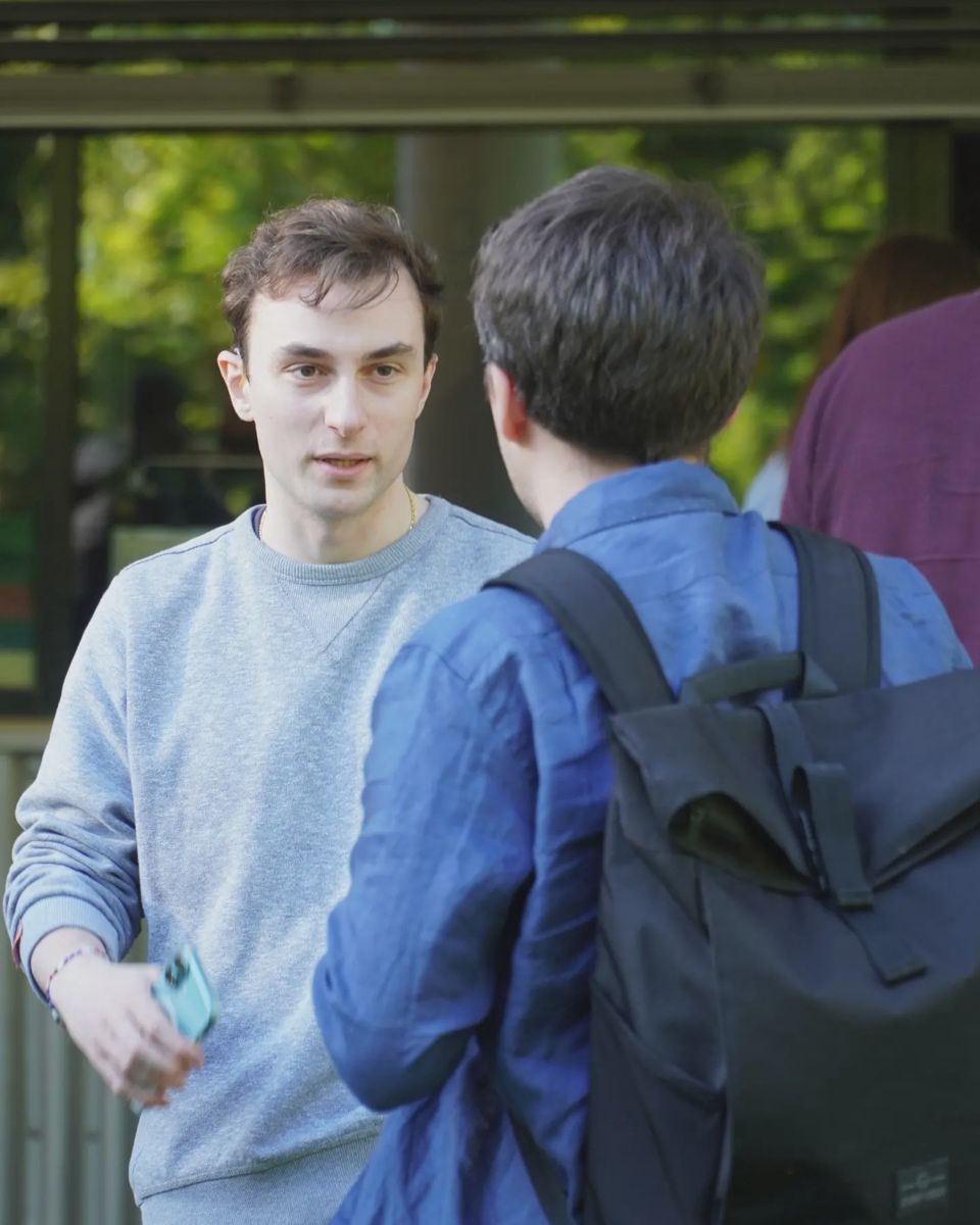 Zwei Personen stehen im Gespräch vor dem Hochschulgebäude; eine trägt einen Rucksack.